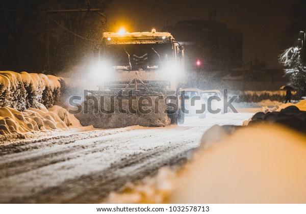 A snow plough mounted on a\
truck is cleaning the road for an early morning commute. Row of\
cars behind a snow cleaning truck on a street in a city at\
night.