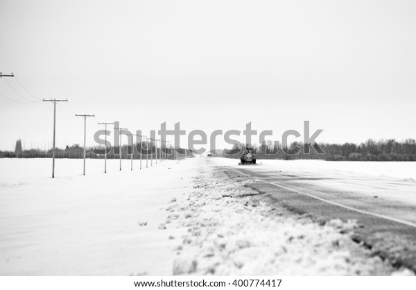 A snow plow clearing a snow laden\
highway in a black and white rural winter\
landscape