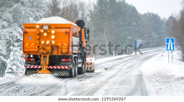 Snow plow on highway salting road. Orange truck\
deicing street. Crystals dropping on snowy asphalt. Maintenance\
winter vehicle in action.