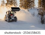 Snow plow tractor cleaning snow with a large bucket on countryside road. Sun backlight. Winter terrain clearing, road safety. Copy space