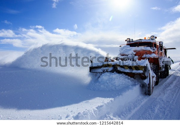Snow plowing truck, huge heavy machine service\
car, snow flakes and splash off road, winter cold snowy season,\
snow cleaning ratrack truck, sunny blue sky day at cold Russian\
arctic north, industrial