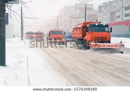 Snow plows remove snow from the road in the city. Several trucks are cleaning the asphalt. It's snowing. Focus blurred.