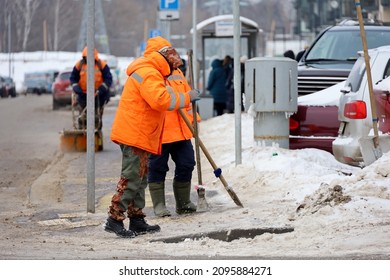 Snow Removal In Winter City, Workers In Uniform Break The Ice With A Crowbars On Sidewalk. Cleaning The Street Near The Cars And Bus Stop