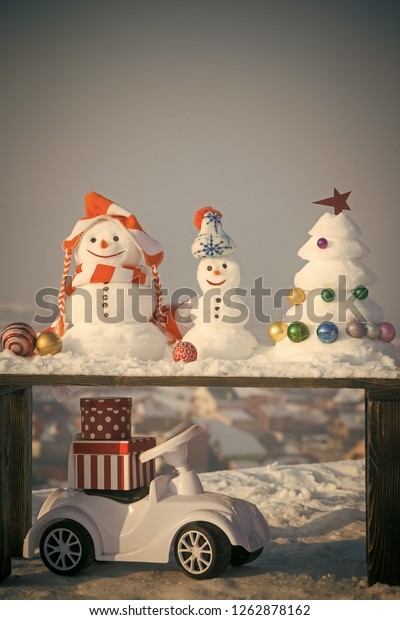 Snow\
sculptures on wooden table on grey sky. Snowmen and xmas tree with\
baubles. Christmas and new year celebration. Toy car with present\
boxes on snowy background. Winter holidays\
concept.