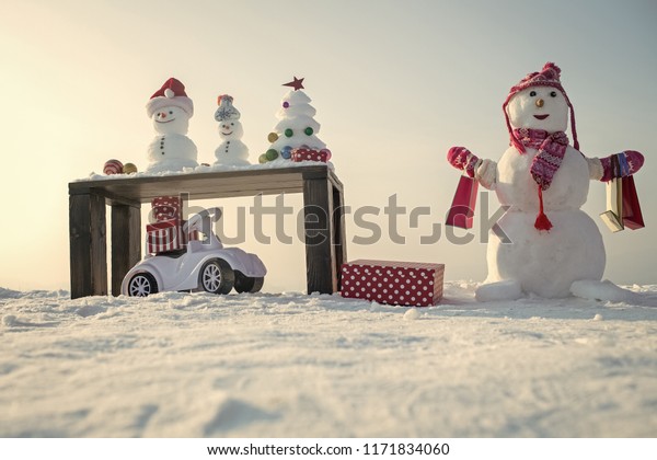 Snow sculptures\
and xmas tree on wooden table. Snowman with shopping bags on snowy\
background. Present boxes and toy car. Christmas and new year.\
Holidays celebration\
concept.