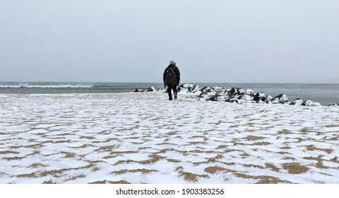 Snow Time On The Coast Of The Sea And Walking Man Through The Beach Nearly To Water. Winter On The Beach Of The Sea.