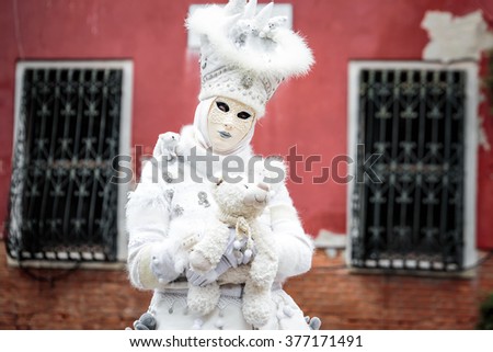 Snow white carnaval mask standing in front red wall background with iron lattice on the window holding toy bear, Venice, Italy.
