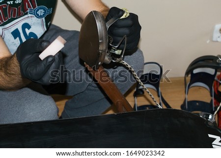 Snowboard service and care. Men's hands in black latex gloves. A man applying wax to the snowboard's slide. Melting wax with an iron and putting a snowboard on the slide. Winter equipment service.