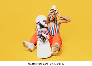 Snowboarder cheerful excited fun woman wear orange ski suit mask hat swimsuit spend extreme weekend sitting hold goggles wink isolated on plain yellow background. Winter sport hobby trip relax concept