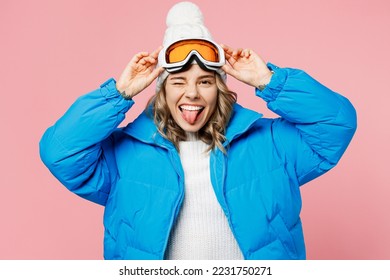 Snowboarder fun woman wear blue suit hat ski padded jacket hold take off goggles mask show tongue wink isolated on plain pastel pink background. Winter extreme sport hobby weekend trip relax concept