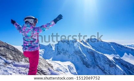 Snowboarder girl with hands raised up in Heiligenblut, Austria. Valley covered with clouds. She wears colourful jacket, sunglasses and helmet. Smiley face shows content and happiness. High Alps around