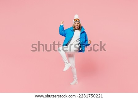 Snowboarder happy woman wear blue suit goggles mask hat ski padded jacket do winner gesture raise up leg isolated on plain pastel pink background. Winter extreme sport hobby weekend trip relax concept
