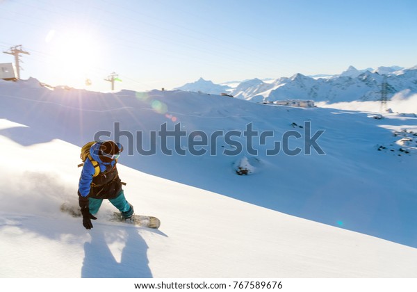 A snowboarder in a ski mask and a
backpack is riding on a snow-covered slope leaving behind a snow
powder against the blue sky and the setting
sun