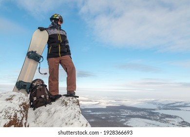 The snowboarder stands on the edge and holds the snowboard. The skier climbed the mountain. Freeride in the mountains. A young man extreme stands with a snowboard on top of a rock.