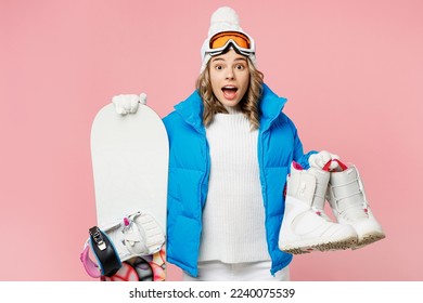 Snowboarder sorprendió a una mujer divertida que lleva traje azul gafas máscara de máscara de máscara de la máscara de esquí que la chaqueta acolchada sostiene botas de snowboard aisladas en un fondo rosa pastel. Concepto de relajación del viaje de fin de semana de hobby deportivo extremo invierno