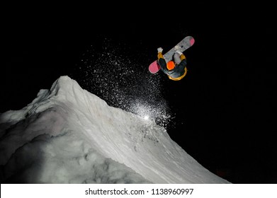 Snowboarder In Winter Outfit With A Snowboard Demonstrates A Beautiful Jump Against The Background Of Snow Mountains At Night