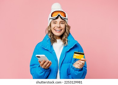 Snowboarder woman wear blue suit goggles mask hat ski jacket use mobile cell phone credit bank card book tour isolated on plain pastel pink background. Winter extreme sport hobby weekend trip concept