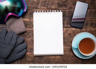 Snowboarding Tips Mock Up. Snowboard Gear List Template. Blank Page Notepad With Copy Space And Snowboard Googles, Cuf Of Black Tea, Mobile Phone On Brown Wooden Table Flat Lay Background.