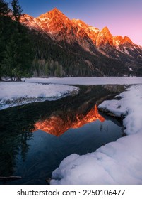Snowcapped Alps mountains reflected warm colors in the partially frozen Anterselva lake (North Italy) at sunset in winter. Snow and ice in the foreground, tree and forest in the background.