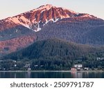 Snow-capped mountain above temperate rainforest and shoreline houses and condominiums on Douglas Island that overlook Gastineau Channel at evening golden hour in mid June. View from Juneau, Alaska.