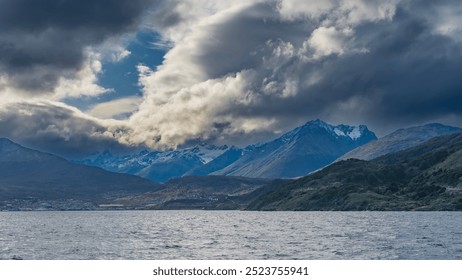 Snow-capped mountain peaks against a blue sky and dark clouds. At the foot of the hills, on the ocean shore, the city buildings of Ushuaia are visible. The Beagle Canal is in the foreground - Powered by Shutterstock