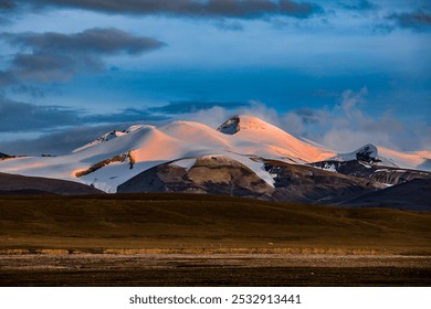 Snow-capped mountains bathed in soft sunset hues beneath a vibrant sky, with a shadowed grassy plain stretching out below. - Powered by Shutterstock