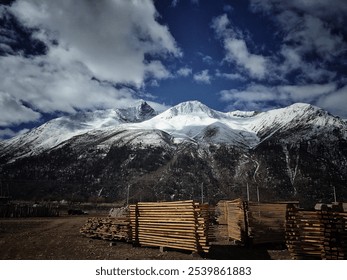 Snow-capped mountains under cloudy sky, stacked wooden planks in foreground, scenic view. - Powered by Shutterstock