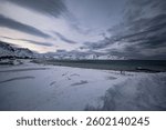A Snow-covered beach with dramatic clouds and mountains in the background, creating a serene winter landscape.