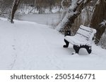 A snow-covered bench sits beside a snowy path near a frozen river, surrounded by bare trees, creating a tranquil and quiet winter scene.