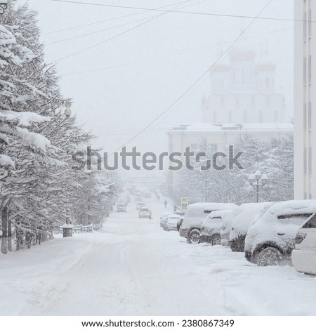 Snow-covered cars parked on the side of a snowy street. Poor visibility during heavy snowfall and snowstorm in the city. Lots of snow on the roadway and trees. Cold winter weather. Magadan, Russia.