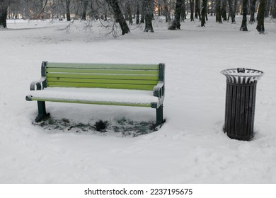 Snow-covered green wooden park bench and black trash can