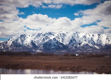 A snow-covered mountain in Logan, Utah sits above a drab Spring farm and field, with a pond reflecting in the foreground.