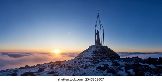 Snow-covered mountain peak at sunrise, featuring monument with statue, cross. Vibrant orange glow of rising sun contrasts with clear blue sky, while sea of clouds stretches out below. Homyak, Ukraine. - Powered by Shutterstock