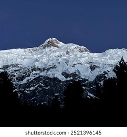 Snow-covered mountain peak under a clear night sky. Layers of glaciers cascade down the rugged slopes, creating a stunning contrast with the dark tree silhouettes in the foreground. - Powered by Shutterstock