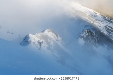 Snow-covered mountain peaks partly obscured by dense fog and clouds. Ski lift structures visible on the slopes - Powered by Shutterstock