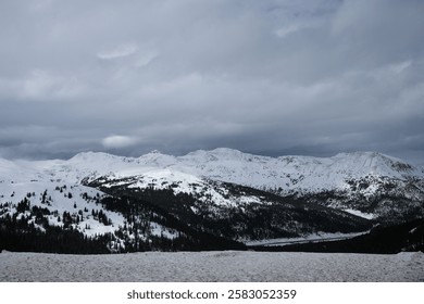 Snow-covered mountain range under dramatic cloudy sky, remote wilderness, winter landscape, rugged alpine terrain, untouched nature, high-altitude scenery, scenic travel destination, moody atmosphere.