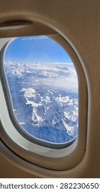 Snow-covered mountain range viewed through an airplane window, with dramatic clouds and clear blue sky creating a breathtaking aerial winter landscape.