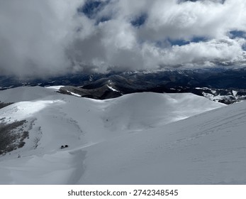 Snow-covered mountain ridge under dramatic clouds with distant valley and solitary cabin