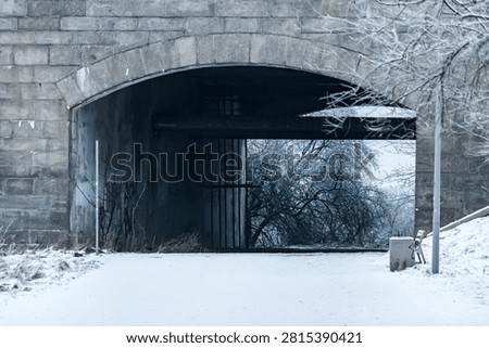 Snow-covered path passing under a stone bridge arch with bare trees and a quiet winter scene
