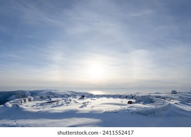 Snow-covered ruins of an abandoned structure on the top of a mountain. There is a solar halo in the sky above the sea. Remains of the foundation of a radiotransparent shelter. Magadan Region, Russia.