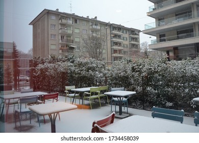 Snow-covered tables and chairs in a tree-lined courtyard, with blocks of flats in the background. Photo taken through a window with atmospheric reflection effects on the left side. 