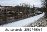 A snow-covered walking path along a river and houses on the other bank on a cloudy winter day, Svartån river in Västerås, Sweden