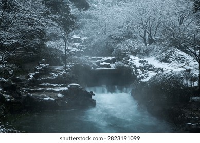 Snow-covered waterfall and stream flow through a quiet winter forest in Taiziwan Park, Hangzhou, creating a serene misty landscape.