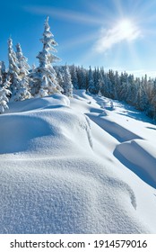 Snowdrifts on winter snow covered mountainside, fir trees on hill top and sun shine in blue sky