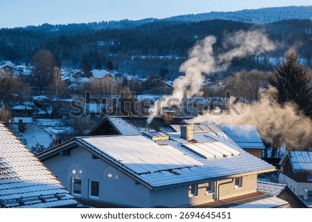Snow-dusted rooftops, solar panels and chimneys sending smoke into cold air above a small German village on a winter morning.