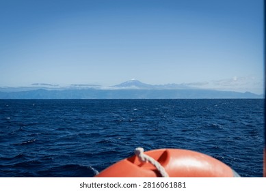 The snow-dusted summit of Mount Teide on Tenerife rises faintly on the horizon across the deep blue Atlantic beyond an orange lifebuoy on the stern of a sailing yacht off La Gomera.