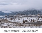 Snow-dusted village along a waterfront, nestled under misty hills, with rocks on the snowy shoreline under a cloudy sky.