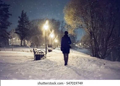 Snowfall In The City Park. Woman Walking Down By Alley. Night Landscape With Falling Snowflakes. Severe Weather In The Winter Park With Trees, Benches Covered By Snow. 