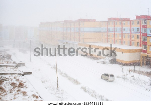 Snowfall in the city. Top view of a snowy city\
street and colorful buildings. Car rides on a road covered with\
snow. Anadyr, Chukotka, Siberia, Far East of Russia. October in the\
Arctic.