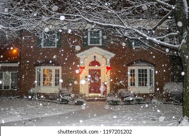 Snowfall On Beautiful Brick House With Columns And Bay Windows With Christmas Tree Light Up And Red Sled And Wreath On Porch